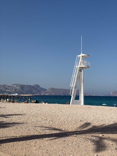 The image depicts a bright, sunny day at a pebble beach in l'Alfàs del Pi, Spain. The prominent feature is a tall, white lifeguard tower with a distinctive spiral staircase and an observation platform. The tower stands near the water's edge, casting a shadow on the pebbled shore. The sea is a clear turquoise, and in the background, a range of hills with scattered buildings and a distant cityscape can be seen under a clear blue sky. Several beach umbrellas and chairs are set up on the beach, and a few people are visible, some in the water and others relaxing on the shore. The foreground of the image is dominated by the textured surface of the pebble beach, with palm tree shadows creating a dappled effect.
