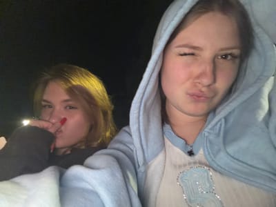 Two young females are captured in a dimly lit, outdoor environment, likely at night. The individual on the right, wearing a light blue hooded sweatshirt over a white top with blue detailing, is winking at the camera with her lips pursed. Her right eye is closed, while her left eye is open. Her hair is a light brown color, and she appears to be wearing makeup.
The female on the left, with light brown hair, is looking towards the camera and has her right hand near her mouth, with her index finger touching her nose. She is wearing a dark colored top and a white garment underneath. Her fingernails are painted red.
The background is dark, suggesting it is nighttime. There are some faint lights visible in the distance, possibly streetlights or car headlights. The overall scene is casual and appears to be a selfie. There is no discernible evidence to suggest the location is in the city of Siedlce, Poland.
