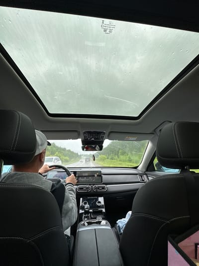 The image shows the interior of a car, looking out through the windshield and sunroof. The weather appears to be overcast and rainy, as indicated by the water droplets on the glass. A person, wearing a grey quilted jacket and a cap, is driving the car. Their hands are on the steering wheel. The car's dashboard and a tablet screen displaying navigation are visible. Outside, the road is lined with green trees and fields, suggesting a rural or suburban setting. Other vehicles are also visible on the road ahead. The time of day is indeterminate due to the overcast conditions. There is a visible safety warning sticker on the car's interior.