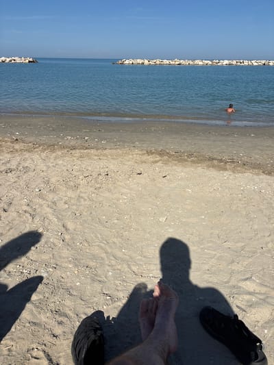 This is a close-up, eye-level, point-of-view shot taken at the beach in Pedaso, Italy. The foreground is dominated by the sandy beach with scattered shells and debris. Two black shoes are visible in the bottom right and left corners, and a person's feet are positioned in the center foreground. Shadows of people are cast on the sand. In the mid-ground, the calm blue water of the Adriatic Sea meets the shore. Two rock breakwaters, constructed of large white stones, extend into the sea on the left and right sides of the frame, creating a sheltered area. In the far mid-ground, a single person, appearing to be a child, is wading in the water. The sky is a clear, bright blue with a faint haze on the horizon.