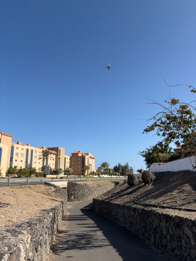 The image shows a clear blue sky with a distant airplane flying in the upper center. Below the sky, a street runs diagonally from the bottom center towards the upper right, bordered by stone walls on both sides. On the left side of the street, there are buildings with light-colored facades and orange roofs, featuring multiple windows. Palm trees and other vegetation are visible around the buildings. The environment appears to be arid or semi-arid, with gravel and dry earth alongside the path. In the foreground, the shadow of trees falls across the street. The scene suggests an outdoor urban or suburban setting with clear weather. The image does not depict any people. The location is identified as Antigua, Spain.
