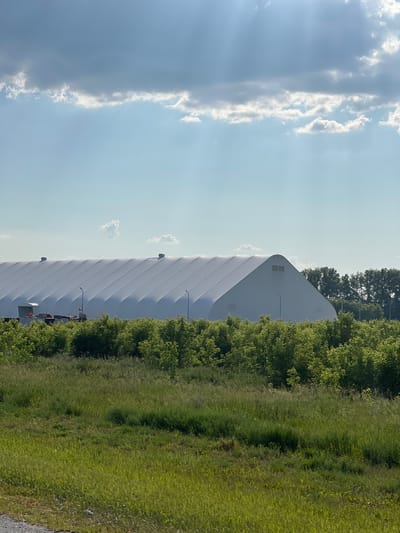 A large, white, arched structure, likely an industrial tent or arena, dominates the midground of the image. It is situated in Verkh-Tula, Russia. The structure is covered with a white, taut fabric that displays a repeating pattern of vertical pleats or segments. Two dark, circular vents are visible on the roof of the structure towards the left side. Towards the left edge of the structure, there are several smaller objects, including a white trailer with an orange tarp on top and what appear to be vehicles. Several light poles are positioned in front of the structure.

The structure is surrounded by dense, green foliage and trees. In the foreground, there is a field of tall, green grass and other vegetation, with some areas appearing more sunlit than others. In the background, behind the white structure, a line of dark green trees forms a backdrop. The sky above is a light blue, with scattered white and gray clouds. Sunlight streams through the clouds, creating visible rays. The overall scene appears to be an outdoor environment on a sunny day. There are no visible people in the image.