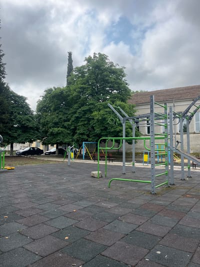 This vertical image depicts an outdoor fitness or playground area in Troyanovo, Bulgaria, under an overcast sky. The central feature is a large, multi-component exercise structure constructed from grey metal tubing with distinctive bright green accents. This equipment includes monkey bars, pull-up bars, and hanging rings. Further in the background, additional fitness apparatuses with yellow and blue elements are visible.
The ground beneath the exercise equipment is covered with a dark, mottled rubber safety surface, consisting of rectangular tiles in varying shades of grey, black, and reddish-brown. To the left and behind the main structure, a large, dense deciduous tree with abundant green foliage stands prominently, with other smaller trees and bushes extending into the distance.
In the background, a light-colored building with a textured facade and dark-tiled roof is visible, partially obscured by the fitness equipment and trees. Parked cars can be seen further back, behind the treeline and some benches. The sky is largely covered with light and dark grey clouds, indicating an overcast daytime. No individuals are present, and no specific activities are taking place. There is no discernible text visible within the image.