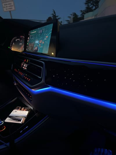 The image shows the interior of a modern car at dusk. The dashboard features a large, wide infotainment screen displaying a map, a digital instrument cluster, and various climate control buttons illuminated with red LEDs. Ambient blue lighting runs along the dashboard and door panel, creating a futuristic ambiance. A smartphone is placed in a charging dock on the center console, displaying a video with a landscape in the foreground and a group of people in the background. The exterior view through the window suggests a location with trees and buildings, possibly on the outskirts of a city, consistent with the general surroundings of Šempeter pri Gorici, Slovenia. The overall scene depicts a comfortable and technologically advanced car interior.