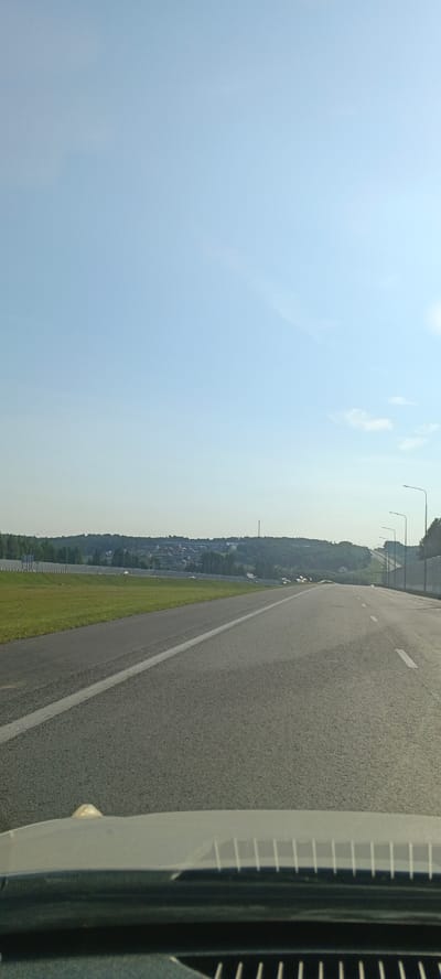 The image shows a view from a vehicle traveling on a paved road. The road has a white line marking the lane. To the left of the road is a green grassy area, followed by a sound barrier and then a further expanse of greenery and trees. In the distance, there are buildings, suggesting a settlement or town, likely Кощаково, Russia, nestled amongst rolling hills covered in trees. The sky is a clear, pale blue with a few wispy clouds. Streetlights are visible along the right side of the road. The foreground shows the dashboard or hood of the vehicle from which the photo was taken, with a reflective surface and what appears to be a grille.