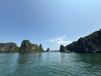 The image depicts a seascape in Tuan Chau Ward, Vietnam, under a bright blue sky with scattered white clouds. The water is dark green, showing ripples across the surface. In the distance, numerous limestone karsts jut out of the water, covered in green vegetation. Several tour boats are visible on the water, some closer to the viewer and others further away between the karsts. The overall scene suggests a popular tourist destination.
