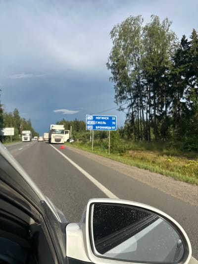 An over-the-shoulder perspective from a vehicle shows a traffic incident on a two-lane highway. The highway appears to be in a rural or suburban area, bordered by trees on the right. A blue directional sign is visible on the right side of the road, indicating distances to Noginsk (33 km), Zhelezhnodorozhny (70 km), and Bronnitsy (97 km), suggesting the location is near Ostankino, Russia. In the distance, two large white trucks are stopped on the highway. The truck in the right lane has its driver's side door open. A red triangular warning reflector is placed on the road behind the white truck. The sky is cloudy with a mix of blue and dark gray clouds. The foreground shows the interior of the vehicle, including a side mirror and part of the car's frame.