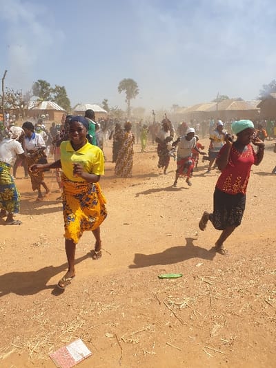 The image depicts an active outdoor scene in Kallah, Nigeria, featuring approximately 30-50 individuals, primarily adult women, engaged in vigorous movement on a dusty, reddish-brown unpaved surface under a bright, hazy sky. Significant atmospheric dust is visibly suspended, particularly in the mid-ground.
In the foreground, two women are prominently positioned in motion. On the left, a young woman wearing a bright yellow short-sleeved top, a patterned yellow, blue, and black knee-length skirt, and open-toed sandals, moves towards the right, smiling directly at the camera. On the right, another young woman, dressed in a red patterned top, dark skirt, and a light blue headscarf, moves rapidly towards the left with her right hand raised to her face. Long, distinct shadows are cast from these figures onto the ground. The immediate foreground ground contains scattered light-colored organic debris, a small green plastic object, and a partially visible red and white patterned booklet.
The mid-ground is densely populated with individuals, mostly women in varied traditional attire, including patterned skirts, tops, and head coverings. Many are in active states of movement, contributing to the visible dust. At least one person in the mid-ground appears to be wearing a white face mask.
The background features several single-story structures with earthen or mud walls and light-colored corrugated metal roofs, consistent with a rural or semi-rural setting. Scattered trees, including a prominent palm tree, are visible. The sky is light blue with sparse, hazy cloud cover, and atmospheric dust obscures distant details.