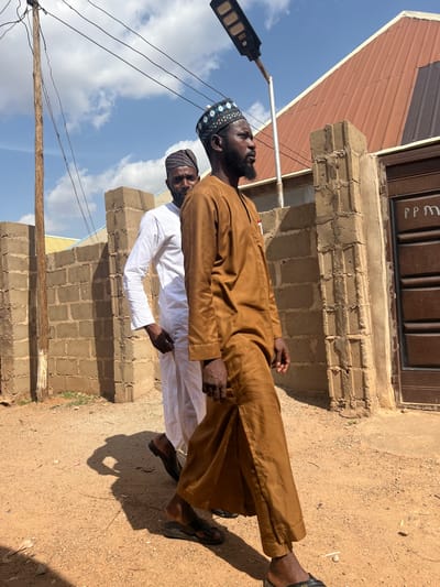 The image shows two men standing outdoors, possibly in the Rigasa area of Nigeria. The man in front wears a brown traditional garment and sandals, with a patterned kufi cap on his head. The second man, partially visible behind him, wears a white garment and a darker kufi cap. A cinder block structure is visible in the background. To the left, there's a building with a reddish-brown roof, and an outdoor light fixture is positioned above the men. The ground appears to be dirt. The sky is partly cloudy.