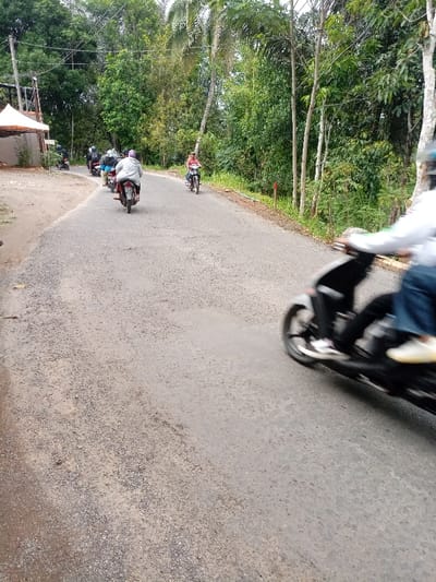 A group of five people are riding motorbikes on a paved road.  The road is in a rural area, with tall trees lining the sides.  One rider is in the foreground and blurred. The other riders are in the background and appear to be driving away from the viewer.  The road is in Kecamatan Cisewu, Indonesia, located at latitude: -7.383, longitude: 107.504.  A white canopy tent is visible off to the left. The road is lightly traveled, and the scene is calm.