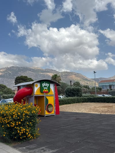 This is a vertically oriented, full outdoor shot of a McDonald's PlayPlace area, set against a backdrop of sun-drenched, rugged mountains under a bright blue sky with scattered white clouds. The play structure is predominantly yellow and red, with a green sign bearing the "play land" logo and the iconic golden arches of McDonald's. A bright red slide curves out from the structure. To the right of the play structure is a vibrant green bush filled with small yellow flowers, and beyond that, a black rubberized playground surface. In the background, a dense, manicured green hedge runs horizontally, with dry, tan-colored ground and more greenery beyond. A building with a reddish roof and glass facade is visible further back, nestled into the hillside. A tall lamppost stands near the building. The overall scene suggests a sunny day, likely midday, in a location that combines urban amenities with a natural, mountainous landscape. There is no visible activity or people in the image. The image appears to be taken in Carini, Italy.