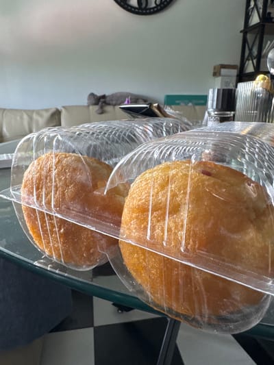 The image shows two round, golden-brown pastries, possibly doughnuts or fritters, encased in clear plastic packaging. The pastries have a slightly textured surface. The packaging itself appears to be a transparent plastic container with horizontal ridging. They are placed on a glass surface, which reflects the checkered pattern of a floor or tabletop in the background. To the left, part of a black and white circular object, possibly a clock face, is visible. In the background, there are hints of household items, including what appears to be a shelf unit with some boxes and a metallic object with a light source. A blurred figure or object, perhaps a stuffed animal, is partially visible behind the pastries on the left. The overall setting seems to be an indoor environment, possibly a kitchen or dining area. There is no visible text in the image. The lighting suggests it might be daytime, but there are no strong indicators of specific time or weather.