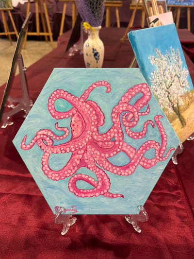 The image displays a hexagonal canvas painting of a pink octopus against a light blue background, positioned on a burgundy tablecloth. The painting is supported by a clear acrylic easel. To the right of the octopus painting, another canvas painting depicting a tree with white blossoms against a light blue sky is visible, also on a stand. In the background, several other paintings are displayed on easels, and a vase with blue floral patterns and a yellow ribbon is present. The environment appears to be an art exhibition or fair, possibly in a public space like a convention center or gallery. The image does not provide specific information to determine the location in Elx / Elche, Spain.