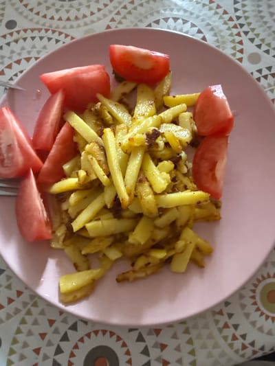 A close-up, high-angle shot shows a pink plate of food, likely a meal in the city of Morozovka, Russia. The plate contains fried potato strips mixed with spices and several wedges of fresh tomato. The food is arranged on a patterned tablecloth with triangular designs in shades of cream, brown, and grey. A fork is partially visible on the left side of the plate. The lighting appears to be natural, coming from above.