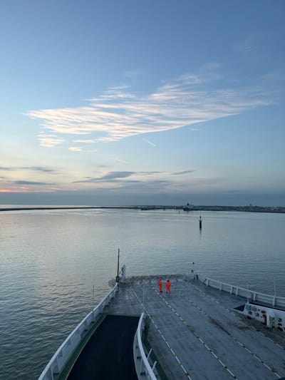 Two individuals wearing bright orange work suits stand on the deck of a ferry in Calais, France. The ferry is docked or moving slowly in a body of water, with a long breakwater extending into the distance. The sky above is a pale blue, with wispy clouds and a contrail from an airplane. In the background, a large ferry or ship is visible, as well as port facilities and buildings along the coastline. The water is calm and reflects the light of the sky. The scene suggests an early morning or late evening setting due to the soft lighting.