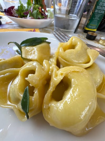 A close-up, elevated view depicts a dining table scene in Anzola dell'Emilia, Italy. The central subject is a white ceramic plate containing multiple servings of light yellow, ring-shaped pasta, consistent with tortellini or tortelloni, coated in a glossy, translucent sauce. Two individual dark green sage leaves are visible, each placed atop a separate pasta piece. A silver four-tine fork rests on the table to the right of the plate. In the background, a small white bowl holds a mixed green salad with red leaf components. Further back and to the right, a clear drinking glass is approximately half-filled with water. Partially visible behind the glass is a dark green bottle labeled "OLIO EXTRA VERGINE DI OLIVA" (Extra Virgin Olive Oil) and a small, gold-colored metallic cap. The entire setting is arranged on a patterned tablecloth, primarily yellow, gold, and white.