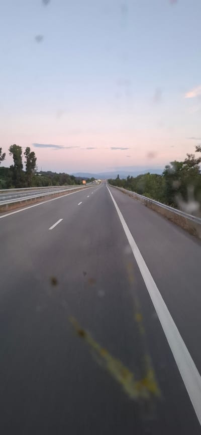 The image shows a forward-facing perspective from a vehicle on a highway at dusk. The sky is a pale gradient of blue and pink, with faint clouds. Trees line both sides of the road, with more dense vegetation in the distance. The highway itself is asphalt with clear white lane markings, and a metal guardrail runs along the left side. In the distance, the highway curves slightly and seems to lead towards a more populated area with lights visible. There are no people or animals visible in the frame, and the scene appears to be quiet and empty, with no other vehicles immediately present. The image quality is slightly affected by what appear to be water spots or smudges on the lens, particularly in the foreground. The general location is described as a highway in Mursalevo, Bulgaria, at the time of twilight.