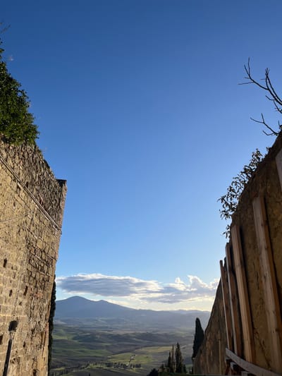 The image displays a panoramic vista of a sunlit landscape and sky, observed from a vantage point framed by ancient architecture. The foreground features weathered stone structures: a robust stone wall partially covered by green ivy or bush in the lower part of the frame, across which a rope or wire runs diagonally; and an architectural overhang or roofline in the upper part, revealing wooden beams and sparse, dried foliage, with bare tree branches extending into the top left sky.

Beyond these architectural elements, a vast expanse of rolling green hills and distant mountains stretches under a clear blue sky. A prominent band of elongated, white clouds is brilliantly illuminated by the low sun, creating a warm, golden glow and a slight atmospheric haze over the distant terrain. The sun's position suggests either early morning or late afternoon.

No individuals or discernible text are present. The scene is devoid of specific activity, capturing a moment of tranquil natural beauty interwoven with historical structures. The rustic architecture and lush landscape are characteristic of the Tuscan region, aligning with the specified location of Pienza, Italy.