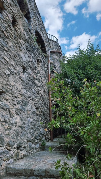 The image depicts a stone wall and a winding staircase leading upwards in Camprodon, Spain. The stone wall is rough-hewn and appears ancient, with small openings suggesting windows or alcoves. A rusty pipe runs vertically along the wall, attached to a building or structure that recedes into the background. Lush green foliage spills over the stone steps and surrounds the base of the wall, obscuring some of the lower portions of the staircase. The sky above is a vibrant blue, dotted with fluffy white clouds, indicating fair weather. A railing is visible on a curved structure at the top of the staircase, hinting at a balcony or viewpoint. The overall scene evokes a sense of history and natural beauty, possibly within the grounds of an old castle or historical building.