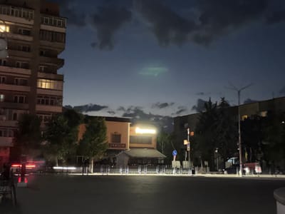 This media file captures a twilight scene in Stryi, Ukraine, likely during the evening as indicated by the dim lighting and illuminated windows of a tall residential building on the left. The sky is a deep blue, fading to lighter tones near the horizon, and is partially obscured by dark, cloudy formations. A peculiar, luminous oblong shape hovers in the sky, possibly an unusual cloud formation or a man-made light.
The foreground features a paved area, likely a street or plaza, with a dark asphalt surface. On the right side, a street lamp with a multi-pronged top stands tall, its light casting a glow on the surroundings. Trees with dense foliage are scattered throughout the scene, some lining the street and others behind buildings.
In the mid-ground, a building with a cream-colored facade is prominently visible, featuring a sign that reads "FAMILIA." Above this, a large, illuminated sign with bright yellow lettering is present, likely indicating a business or establishment. A smaller, darker structure with a slanted roof is situated in front of the "FAMILIA" building. Metal barriers or fences are placed in front of these buildings.
Further to the right, another building, partially obscured by trees and darkness, is visible. Vehicles, including what appears to be a dark-colored car and a red van or truck, are parked or driving in the background. The overall atmosphere is one of quiet evening activity in an urban or semi-urban environment. The presence of lit windows and illuminated signs suggests that life continues despite the approaching night. The faint red light streaks on the left hint at passing vehicle lights.