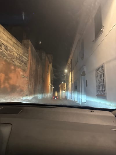 The image depicts a nighttime view from the interior of a vehicle traveling through a narrow street in Orosei, Italy. The street is flanked by old buildings with weathered facades, some painted in muted oranges and others in white. Streetlights illuminate the scene, casting a yellowish glow on the left side and a cooler light on the right. There is evidence of moisture or condensation on the windshield, creating a slightly diffused and hazy effect. In the distance, the taillights of another vehicle are visible, indicating movement along the street. The architecture suggests a historic Mediterranean town. The perspective is low, as if taken from the driver's or front passenger's seat, with the dashboard visible in the foreground. The sky above is dark, typical of nighttime.