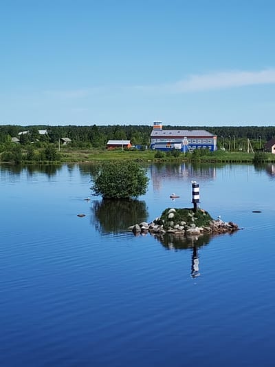 This image depicts a serene summer landscape in Shestovo, Russia, characterized by a calm body of water under a bright blue sky.
In the foreground, a small, rocky islet emerges from the water, topped with green moss and featuring a miniature black and white striped lighthouse. Several white birds, likely gulls, are perched on the rocks and floating in the water nearby. To the left of the islet, a dense, spherical green bush grows directly from the water. The tranquil water surface reflects the sky and the objects on its surface, including the lighthouse and the distant buildings.
The midground reveals a lush, green shoreline densely covered with trees, forming a forest line along the horizon. Several buildings are visible along this shore. A prominent blue and gray building with an orange-topped tower stands out, featuring multiple windows and solar panels on its roof, suggesting a modern facility. To the left, several smaller residential-style buildings, including one with a red roof, are nestled amongst the trees. Utility poles and wires are also visible along the shore.
The time of day appears to be bright daylight, possibly midday or early afternoon, given the high sun and clear, cloudless sky. The overall scene conveys a sense of peace and tranquility, combining natural elements with distant human infrastructure. No discernible text is visible.