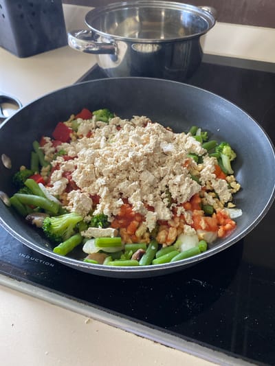 The image depicts a black frying pan on a black induction stovetop, likely in a kitchen. The pan contains a mixture of vegetables and what appears to be tofu. The vegetables include broccoli, green beans, red peppers, mushrooms, and onions. Tomato sauce is also visible. The tofu is crumbled and appears to be the main protein source in the dish. To the left of the frying pan is a stainless-steel pot with its lid off. The stovetop is on a light-colored countertop. The scene appears to be in a home kitchen. There is no indication of the specific city of Villaviciosa, Spain.