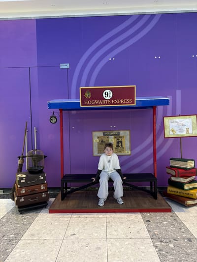 The image depicts a staged scene resembling Platform 9 3/4 from the Harry Potter series. A young boy is sitting on a luggage cart, dressed in a white top and grey pants. Behind him is a purple wall with the "Hogwarts Express" sign prominently displayed above. Additional elements include a framed poster with a Daily Prophet headline about Harry Potter, a framed certificate, a stack of books, and a pile of vintage suitcases with a birdcage on top. The setting appears to be indoors, with a tiled floor in the bottom right corner of the image. The specific location within Родниковское сельское поселение, Ukraine, cannot be definitively determined from the visual information.