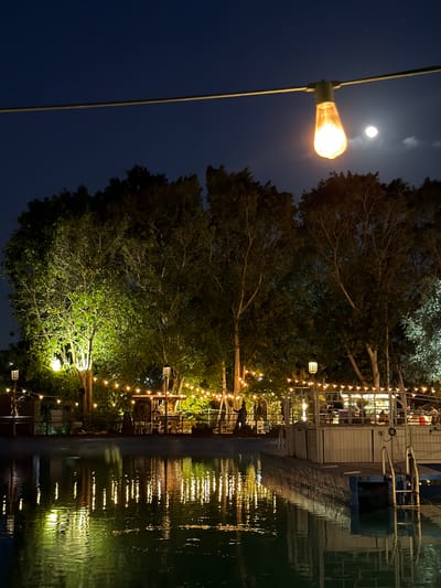 When correctly oriented, this nighttime photograph from Nir Eliyahu, Israel, captures a serene outdoor scene, likely a restaurant or cafe, situated alongside a calm body of water. The dark blue sky, indicating deep night, features a prominent, nearly full moon glowing brightly, accompanied by a tiny, distant white speck, possibly a star or planet.
In the upper left foreground, a single, glowing Edison-style light bulb hangs from a wire, casting a warm, orange light. Below it and extending into the midground, several large trees with dense, dark foliage are visible. Some parts of the trees' leaves are illuminated by the ambient and decorative lights, showing vibrant green patches.
Numerous warm-toned string lights are strung across the scene, creating an inviting and festive atmosphere. These lights lead towards a building on the right side of the image, which appears to be a restaurant or bar. Windows reveal interior lights and vague silhouettes, suggesting people are gathered inside. A structure, possibly a dock or walkway, runs along the edge of the building next to the water.
The body of water, occupying the lower and right sections of the image, beautifully reflects the string lights and the building's illumination, creating shimmering, elongated streaks of light on its surface. The overall impression is one of tranquility and coziness. There is no visible text in the image.
