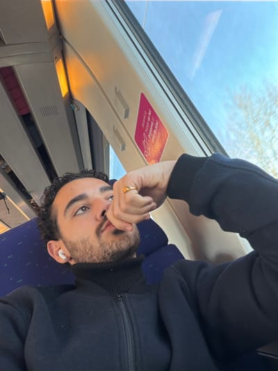 The image displays a male individual seated inside a train carriage, identified by context as being in the city of Chauny, France. The subject has short, dark, curly hair, a beard, and a mustache. He wears a black high-neck zippered garment. A white earbud is inserted into his left ear. His right hand is raised, with fingers positioned near his mouth, and a gold-colored ring is visible on the ring finger of this hand. He is looking upwards and to his right. The train interior includes blue patterned seats and white overhead panels with some illuminated orange-yellow light fixtures. A pink rectangular sticker with Dutch text, "Bedankje op het dagmenu of onze de zetel!", is affixed to the overhead panel near the window. Outside the window on the right, a clear blue sky and bare tree branches are visible, indicating daytime and a season without foliage. The background elements appear slightly blurred, consistent with the movement of the train.