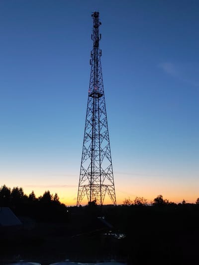 A low-angle shot depicts a tall metal lattice telecommunications tower silhouetted against a gradient sunset sky. The sky transitions from deep blue at the top to shades of orange and yellow near the horizon. The tower extends high into the frame, with antennas and other equipment visible near the top. The base of the tower is situated behind a dark treeline and a building.
The image was taken in Annensky Most, Russia.