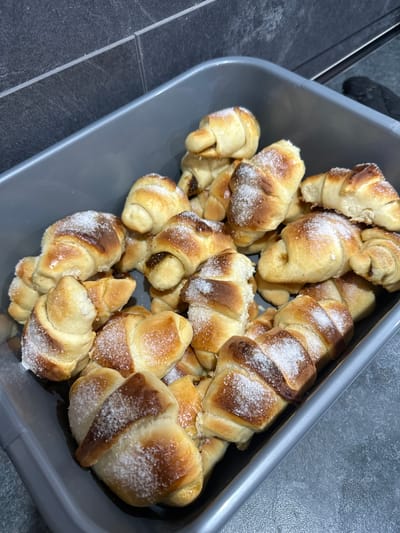 A gray plastic container filled with crescent-shaped pastries, sprinkled with white sugar, sits on a dark gray tiled surface. The location in Vinarovo, Bulgaria cannot be determined from the image.