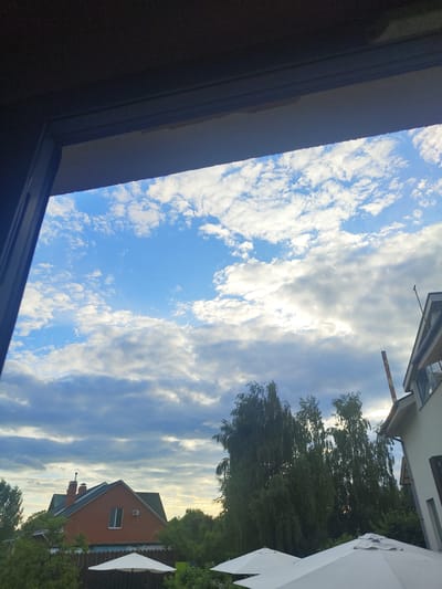 The image depicts an outdoor scene viewed through a window frame, looking out towards a residential area under a cloudy blue sky. In the foreground, partially obscuring the view, are the dark, textured surfaces of the window frame, likely made of wood or a similar material.
Beyond the window, the sky is predominantly blue with scattered white and gray clouds. The clouds vary in density, with some appearing wispy and others more substantial, suggesting a dynamic weather pattern. Sunlight is visible breaking through some of the cloud cover, creating brighter areas in the sky.
In the mid-ground, several residential buildings and trees are visible. To the left, a two-story house with a red brick exterior and a dark pitched roof is prominent. It features a chimney and a single visible window. Surrounding this house and extending across the scene are lush green trees, some of which are tall and leafy, possibly birch trees due to their slender appearance.
To the right of the frame, a portion of another building, possibly a two-story house with a light-colored facade and a dark roof, is visible. A tall, cylindrical structure, resembling a chimney or a pipe with a reddish-brown hue and white bands, extends upwards behind the trees.
In the foreground, at the bottom of the image, two white umbrella-like structures are visible, likely patio umbrellas, suggesting an outdoor seating or recreational area.
The scene appears to be set in a suburban or rural residential area, consistent with the location of Цибино, Russia. The overall impression is a pleasant, possibly late afternoon or early evening, view from indoors looking out at a tranquil outdoor environment. There are no people or discernible actions captured in the image.