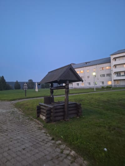 The image depicts a well in a park-like setting, with a building visible in the background. The well is constructed of dark wood logs and has a wooden roof. A bucket is attached to a chain hanging from the well's mechanism. The area around the well is covered in green grass, with a paved walkway in the foreground. In the background, a multi-story building with lit windows suggests a residential or institutional purpose. The sky is a clear blue, indicating it is daytime. A signpost with text is partially visible to the left of the well. The overall scene appears to be calm and orderly.