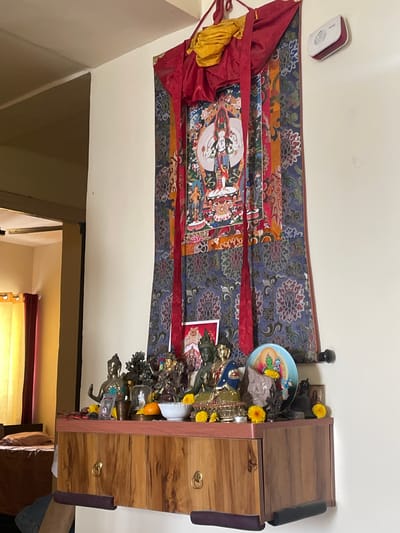 This image displays an indoor religious altar or shrine, likely situated in a home in Noida, India. The central focus is a vibrant thangka painting depicting a multi-armed Buddhist deity, possibly Tara, adorned with intricate details and surrounded by colorful patterns. This thangka is prominently displayed against a light-colored wall and is framed by decorative fabric banners in shades of red, blue, and gold, featuring floral and geometric motifs.
Below the thangka, on the top of a wooden cabinet or shelf unit, numerous religious artifacts are arranged. These include various metallic statues of Buddhas and bodhisattvas in different poses, along with offerings such as fresh yellow flowers (possibly marigolds), a piece of orange fruit, and other items placed within a blue, patterned bowl. The wooden cabinet itself features two drawers with circular golden handles and has a distinctive wood grain pattern.
A small, white, rectangular device, possibly a smoke detector or an alarm, is mounted on the wall to the upper left of the thangka. The arrangement suggests a dedicated space for spiritual practice, prayer, and devotion. The image is taken from a slightly elevated side angle, with the setup extending from the wall onto the wooden unit.