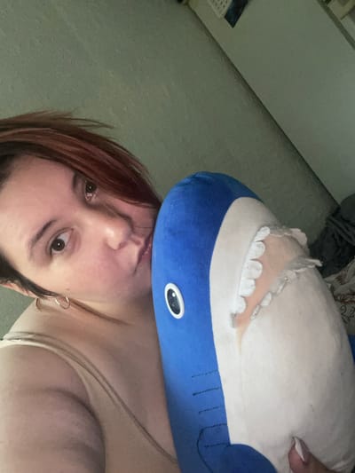 A close-up, overhead, selfie-style shot shows a person holding a stuffed blue shark. The person has fair skin, short, reddish-brown hair, and is wearing a light-colored camisole. Their left eye is visible and open, while their right eye is partially obscured. Their lips are pursed and appear to be kissing the shark. The shark has white felt teeth and a pinkish inner mouth. The background is a light green, textured wall. The image appears to have been taken indoors, possibly in a bedroom, given the visible bed linens in the upper right corner and a calendar on the wall. There is no visible indication of the specific location in Коробицыно, Russia.