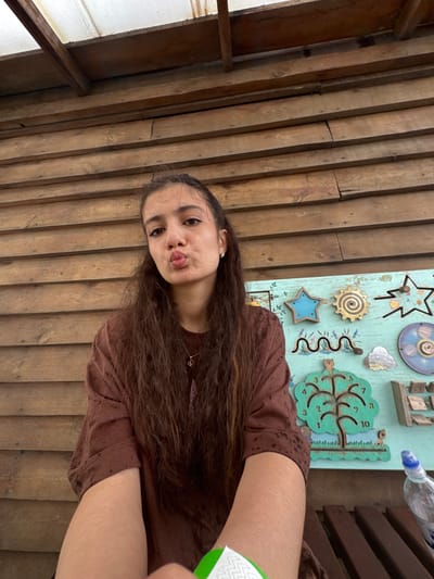 A young woman with long, dark hair is sitting in front of a wooden wall in Zaliznychne, Ukraine. She is wearing a brown shirt and making a kissing face at the camera. To her right is a colorful sensory board with various interactive elements, including gears, stars, and a wooden tree with numbers. The sensory board appears to be attached to the wooden wall. In the foreground, a plastic bottle of water is visible. The lighting suggests it is daytime, possibly outdoors or in a well-lit indoor space with natural light. The background consists of horizontal wooden planks forming a wall.