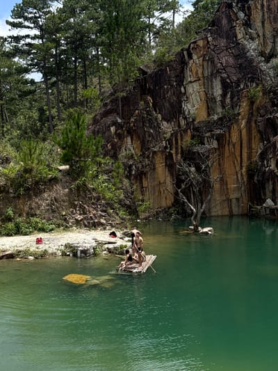 The image depicts a quarry lake in Lán Tranh, Vietnam. A group of people are on a bamboo raft in the emerald green water. The raft is positioned in the foreground to the left of a large, imposing rock face that dominates the background. The rock face is striated with various colors, including brown, tan, and grey, indicating its geological composition. Lush green vegetation, including pine trees, grows along the top of the quarry and on the slopes leading down to the water. The water itself is clear and calm, with visible ripples on the surface. On the shore to the left, there is a red object, possibly a life vest, and some bags. The overall scene suggests a recreational activity in a natural, rugged environment.