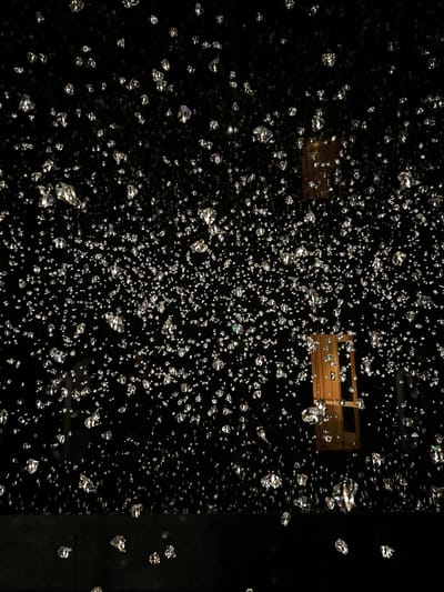 In Wattens, Austria, an art installation features thousands of suspended, multifaceted crystal-like objects against a dark background. These objects, resembling diamonds or ice shards, are illuminated and reflect light, creating a shimmering, starry effect. The crystals vary in size and are densely scattered, with some appearing closer to the viewer than others. In the mid-ground, a wooden structure, possibly part of the installation or an architectural element, is visible. The overall impression is one of depth and sparkle, a seemingly endless field of light-catching fragments. The bottom of the image shows a dark, reflective surface, potentially a floor or a platform. There are no people visible in the scene.