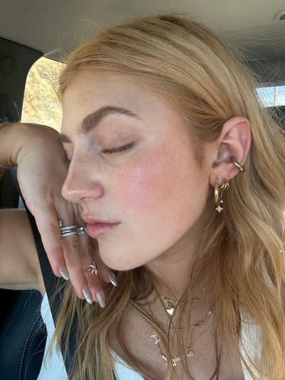 A woman with light blonde hair is shown in a close-up, resting her head on her hand. Her eyes are closed and she has light skin with pink undertones. She is wearing multiple gold earrings, including a cuff, rings, and a necklace with letter charms. She is also wearing several rings on her fingers. Her nails are long and have a metallic silver polish. The photo appears to be taken inside a car.