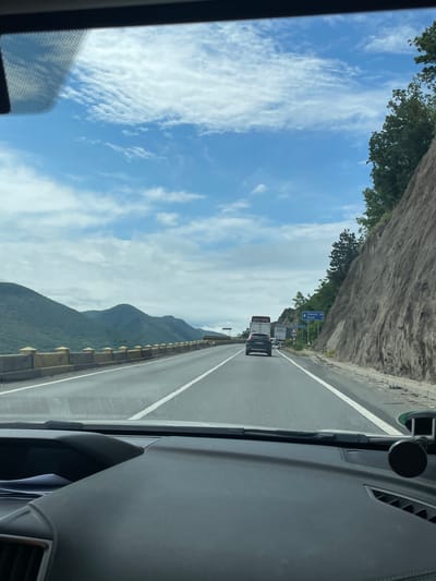 The image displays a daytime view from inside a vehicle traveling on a highway through a mountainous region. The perspective is looking forward through the windshield, with parts of the dashboard visible in the foreground on the right, including vents and a mounted device.
The highway features at least two lanes in the direction of travel and appears well-maintained. On the left side of the road, a steep, rocky cliff face is covered with lush green trees. To the right, a mountain range extends into the distance, with a concrete guardrail lining the highway's edge. The sky is bright blue, dotted with scattered white, wispy clouds.
Several vehicles are visible ahead on the highway, including a large white truck followed by a dark-colored SUV or car. A prominent blue road sign on the left of the highway provides directional information, clearly stating: "↑ Turnu-Severin 10km" and "Orșova." Below these, the text "Portile de Fier I" (Iron Gates I) is partially visible. These location cues, combined with the context, indicate the scene is in Romania, near Drobeta-Turnu Severin.