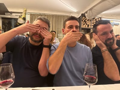 Three men are seated at a table in what appears to be a restaurant. Each man is demonstrating a different action: the man on the left covers his eyes with his hands, the man in the center covers his mouth, and the man on the right covers his ears. In front of each man is a wine glass filled with red wine. A champagne bottle balloon is visible in the background, along with other people dining.