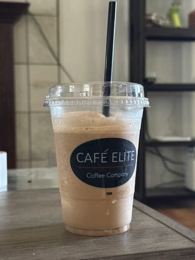 A close-up, eye-level, outdoor shot captures a plastic cup filled with a blended, light brown beverage, likely a coffee drink, on a wooden table. A black straw extends from the cup's clear plastic lid. The cup features a black oval sticker with white text that reads "CAFÉ ELITE Coffee Company" and "TKR" at the bottom. The background is blurred, showing a wall with a tiled pattern and a dark shelf unit. The lighting is natural, suggesting it's daytime. There are no people or animals present. The scene is indoors, but the presence of a wooden table and the natural lighting suggest it might be a cafe or a home with a view of the outdoors. The location is identified as Spanaway, United States of America, but no specific details about the city are visible in the image itself.