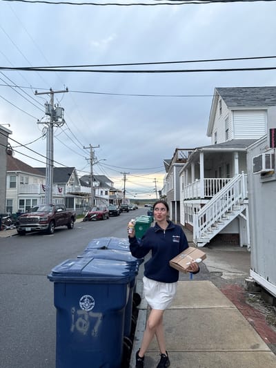A street scene in Hampton, New Hampshire, USA, shows a young woman standing next to blue recycling bins. She's holding a small cardboard box with a white plastic spoon stuck on it and another item in her other hand. She's wearing a navy blue top with a logo, white shorts, and black sneakers. The street is lined with houses, telephone poles, and parked cars. The sky is overcast.