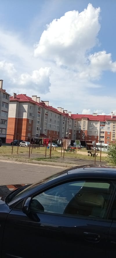 The image depicts a residential area in Hantsavichy, Belarus, characterized by multi-story apartment buildings with red roofs. In the foreground, a dark-colored car is partially visible, taking up the bottom portion of the frame. Behind the car, a paved road leads to a grassy area where clothes are hung on a clothesline. Several cars are parked in the background, including a silver sedan and a black car. A white bus is also visible further in the distance. The sky is blue with scattered white clouds. The scene appears to be an ordinary day in a suburban or urban environment.