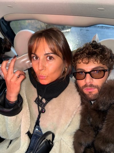A selfie features two individuals in what appears to be a car. The woman in the foreground has dark brown hair with bangs and wears a light, shaggy coat over a black hoodie. Her face is angled towards the camera, displaying brown eyes and a neutral expression. Behind her is a man with curly brown hair, glasses, and a dark fur coat. The car's interior, including a headrest and window with rain streaks, is visible in the background.