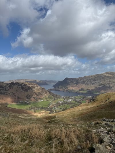 The image displays an elevated panoramic view of the Glenridding valley and Ullswater lake, located in Glenridding, Cumbria, United Kingdom. The foreground features a steep, grassy slope covered in dry, light brown and green grasses, with scattered rocks and a descending dirt path. In the midground, the village of Glenridding is visible, composed of a cluster of white-colored buildings and structures nestled within verdant green fields and interspersed trees. Adjacent to the village, the dark blue surface of Ullswater lake extends, with several small, white objects on its surface, consistent with boats. The lake and village are framed by undulating mountains and fells, displaying a mosaic of brown, ochre, and green vegetation, with areas of exposed rock. The sky occupies the upper portion of the frame, characterized by a mix of clear blue patches and extensive white and grey stratocumulus clouds, casting dynamic shadows across the landscape. No individuals or direct actions are depicted.