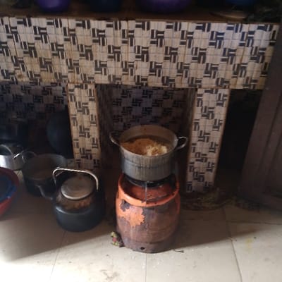 The scene is a kitchen in Adanisievbe, Nigeria, located at coordinates 5.953 latitude and 5.705 longitude. A pot containing food is being heated on a gas burner. The gas burner is mounted atop a large, rusted, orange cylinder. Three additional pots are visible in the foreground. One of these pots is black and is in the lower right corner. The second pot is black and is to the left of the first. The third pot is silver and is behind the second pot and to the left. The kitchen counter behind the burner is constructed of a white and black tiled pattern. A blue bucket is visible to the left of the black pot. The floor is a light brown tile. It is difficult to ascertain if the scene is indoors or outdoors as the walls are tiled and the floor is tile. The kitchen is located at a residence. The scene is likely from a rural area. The objects and the environment are consistent with poverty.