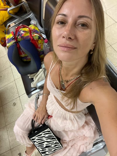 The image shows a woman sitting on a bench, likely in an airport waiting area, given the presence of luggage. She is taking a selfie, and her face is close to the camera, with her blonde hair falling around her. She is wearing a light pink, ruffled dress and several necklaces. She holds a black and white striped bag. Her facial expression appears neutral.
Behind her, several pieces of luggage are visible, including a colorful patterned backpack and a blue neck pillow. A clear plastic bottle of water is also on the bench. The flooring appears to be tiled. The lighting suggests it is daytime. The overall atmosphere is relaxed, indicating she is likely waiting for a flight. The location context of Katunayake, Sri Lanka, implies she may be at Bandaranaike International Airport.
