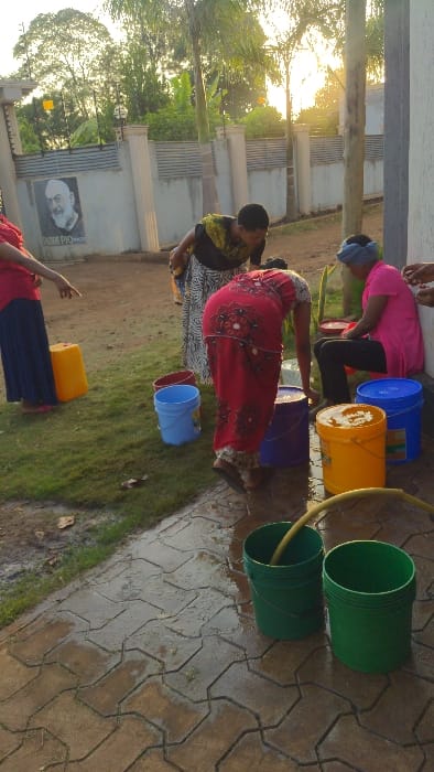 Several people are filling colorful buckets with water from a yellow hose. The buckets are various colors including green, blue, purple, and yellow. They stand on a paved surface that is wet. In the background, there is a building with a portrait on its wall. There are trees and the sun is shining.